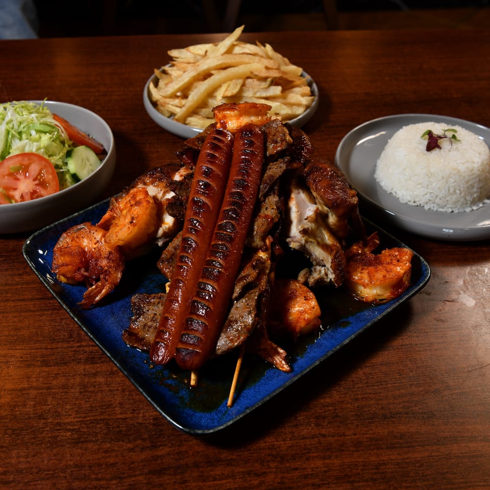Mixed grilled meats, sausages, and shrimp served with white rice, fries, and a fresh salad on a wooden restaurant table.