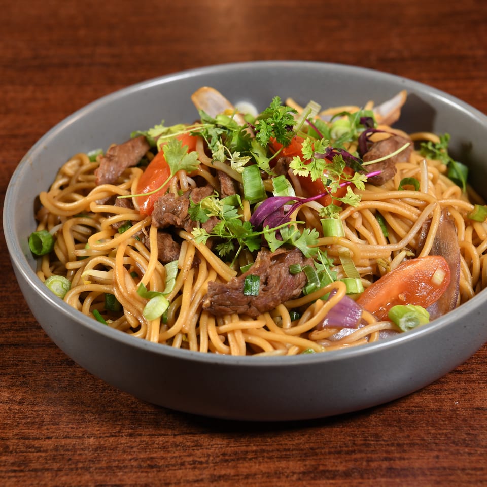 Bowl of stir-fried noodles with sliced beef, tomatoes, onions, scallions, and fresh herbs on a wooden table.