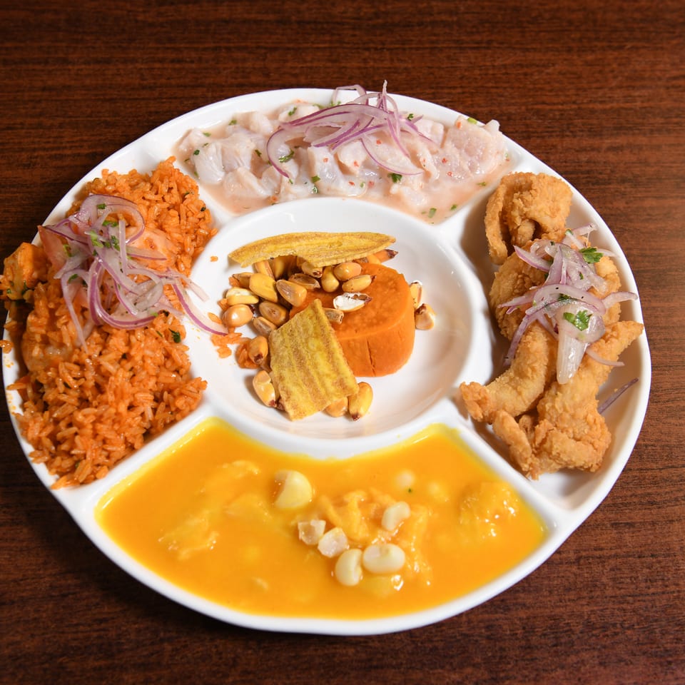 Peruvian sampler platter with ceviche, seasoned rice, fried seafood, plantains, corn, and creamy orange sauce on a wooden table.