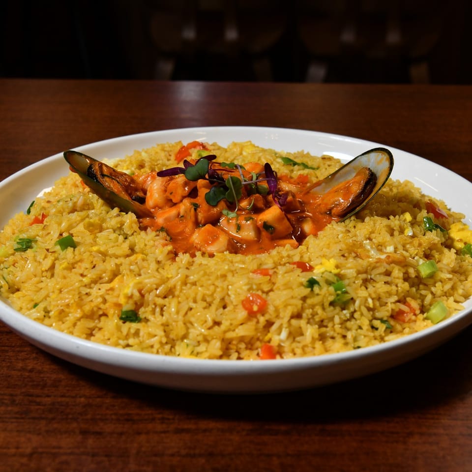 A white bowl of yellow fried rice with seafood in a red sauce and two mussels.